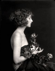 an old black and white photo of a woman with flowers in her hair holding a vase