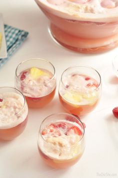 four glasses filled with fruit and ice cream on top of a white table next to a bowl of strawberries