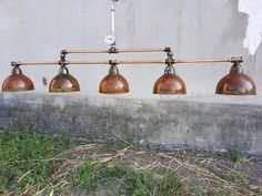 four copper colored lights hanging from a metal bar in front of a concrete wall with grass and weeds