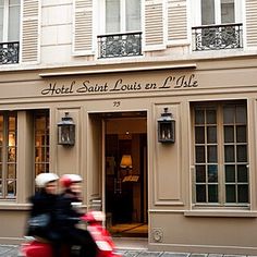 two people on a red scooter in front of a hotel with white shutters