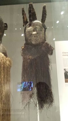 two african masks are on display in a glass case, one is wearing grass and the other has long hair