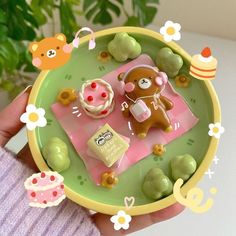 a hand holding a small toy bear and cupcakes on a plate with flowers in the background