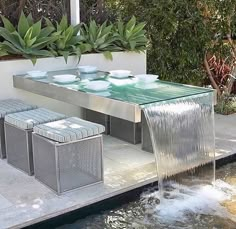 a glass table with water running from it to the ground in front of some plants