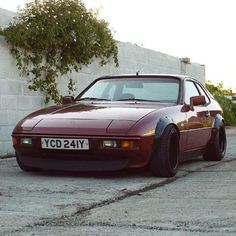 a maroon sports car parked in front of a white brick wall and green bushbery