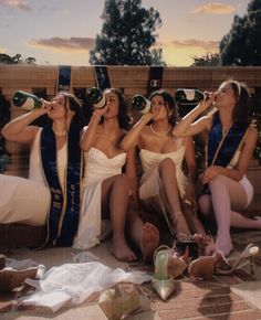 four women in bathing suits sitting on the ground drinking from wine bottles and looking up into the sky