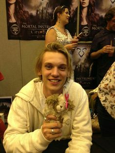 a young man holding a stuffed animal in front of a group of people with posters on the wall behind him