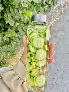 a person holding up a glass bottle filled with cucumbers in front of some bushes