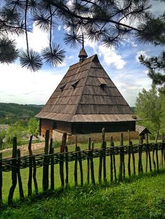 a wooden house with a thatched roof and fence around it's perimeter, surrounded by pine trees