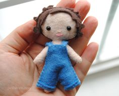 a hand holding a tiny doll with brown hair and blue overalls, in front of a window