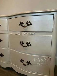 a white dresser with black handles and ornate carvings on the bottom drawer, in a bedroom