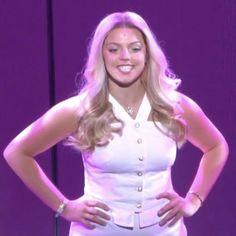 a woman standing in front of a purple background wearing a white dress and gold bracelets