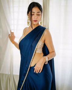 a woman in a blue sari with gold accents on her chest and arms, standing next to a window