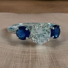 a three stone ring with blue and white stones on the sides, sitting on a piece of cloth