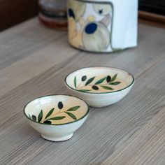 two white bowls with olives painted on them sitting on a wooden table next to a napkin dispenser