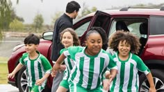 children in green and white soccer uniforms running towards a red car on the side walk