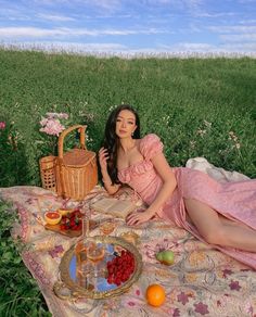 a woman in a pink dress sitting on a blanket next to a basket and fruit