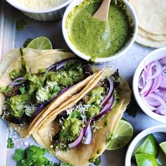 three tacos with green sauce, onions and cilantro on a tray next to tortillas