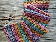 a multicolored crocheted scarf is laying on a wooden surface next to a piece of wood