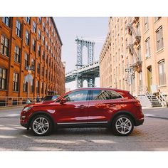 a red car parked on the side of a street next to tall buildings with a bridge in the background