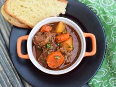 a black plate topped with a bowl of stew next to a slice of bread on top of a green table cloth