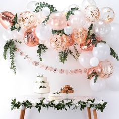 a table topped with lots of balloons and greenery next to a cake on a plate