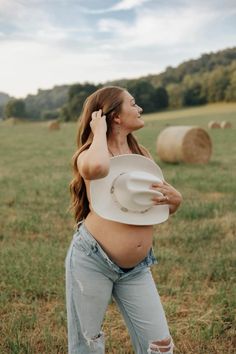 a pregnant woman standing in a field with hay bales behind her back to the camera