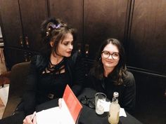 two women sitting at a table with drinks and papers in front of them, both smiling