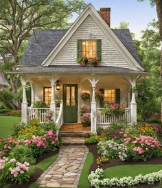 a white house with green shutters and flowers on the front porch, surrounded by greenery