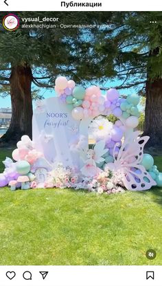 a decorated sign with balloons and flowers on the grass in front of two large trees