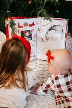 two children are sitting on the floor in front of a christmas tree and looking at an open book