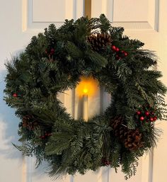 a christmas wreath with a lit candle in the center and pine cones around it, hanging on a white door