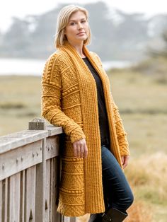 a woman leaning against a wooden fence wearing a yellow cardigan and black top with boots