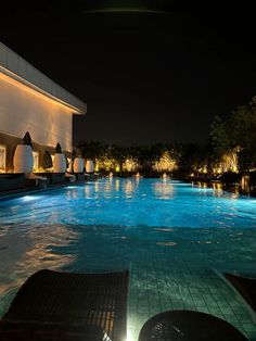 an empty swimming pool at night with lights on the side and lounge chairs around it