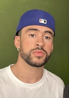 a man with a beard wearing a blue hat and white t - shirt looking at the camera