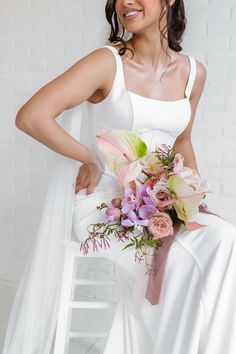 A Bride Sitting In A White Chair With A White Background, The Bride Is Looking To The Side Smiling Holding Her Bridal Bouquet.