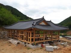 a wooden building sitting on top of a dirt field next to a mountain covered in trees