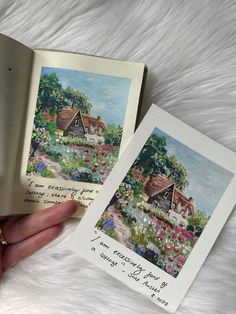 an open book with two pictures of houses and flowers on the pages, in someone's hand