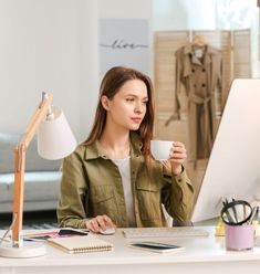 a woman sitting at a desk with a cup in her hand and looking at the computer screen