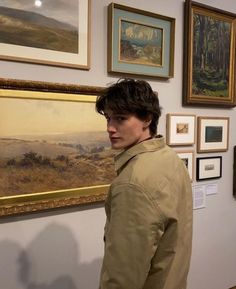 a man standing in front of a wall with paintings on it and framed pictures behind him