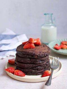 a stack of chocolate pancakes on a plate with strawberries and milk in the background