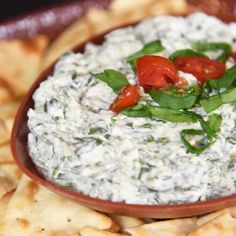 a bowl filled with dip surrounded by crackers and tomatoes on the side, topped with basil