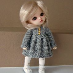 a doll with blonde hair wearing a gray knitted dress and white shoes is standing in front of a wall