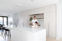 two people standing in a kitchen with white cabinets and counter tops, while one person stands at the island