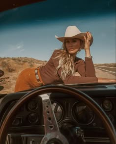 a woman sitting in the drivers seat of a car wearing a cowboy hat and holding her hand up to her head