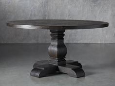 a round wooden table sitting on top of a cement floor next to a gray wall