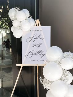 a welcome sign with white balloons in front of a black and white door at an event