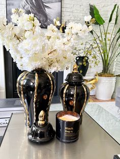 two black vases with white flowers in them on a table next to a candle