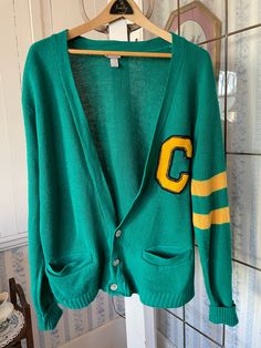 "This classic sweater is made of 100 per cent acrylic in emerald green, with two stripes on the left sleeve in golden yellow. The monogrammed \"C\" is made from thick cotton in the matching yellow and trimmed with black. The cardigan has buttons in the front and two front pockets. Marked size large. The measurements, taken with the sweater lying flat, are: shoulder to shoulder, 22 inches; armpit to armpit, 25 inches; sleeves (uncuffed), 28 inches; length, 25 inches; bottom edge, 22 inches (with stretchy ribbing). The sweater overall has quite a bit of stretch. In good vintage condition." Winter Varsity Cardigan, Varsity Cardigan For Winter College Season, Varsity Style Winter Cardigan For College, Varsity Style Cardigan For College In Winter, Green Cotton Varsity Jacket, Varsity Cardigan With Ribbed Cuffs, Collegiate Cotton Long Sleeve Cardigan, Collegiate Long Sleeve Cotton Cardigan, Classic Green Cotton Cardigan