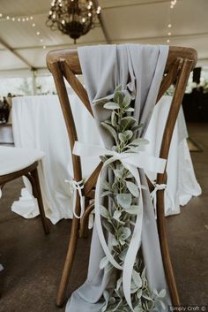 the back of a chair is adorned with white ribbon and greenery as it sits under a tented ceiling