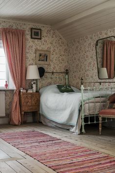 a bedroom with floral wallpaper and pink curtains on the windowsill, a bed in an old - fashioned room